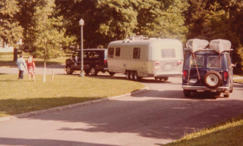 A van with two canoes strapped to the top follows another van pulling a camping trailer